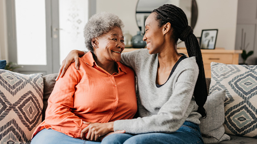 Mother and daughter on couch with arms around each other smiling at each other.
