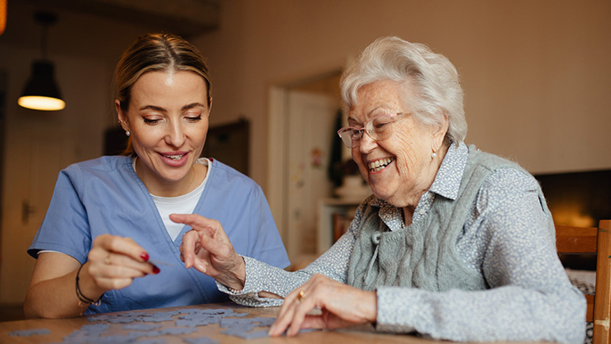 Elderly woman smiling while working on a puzzle with a young nurse.
