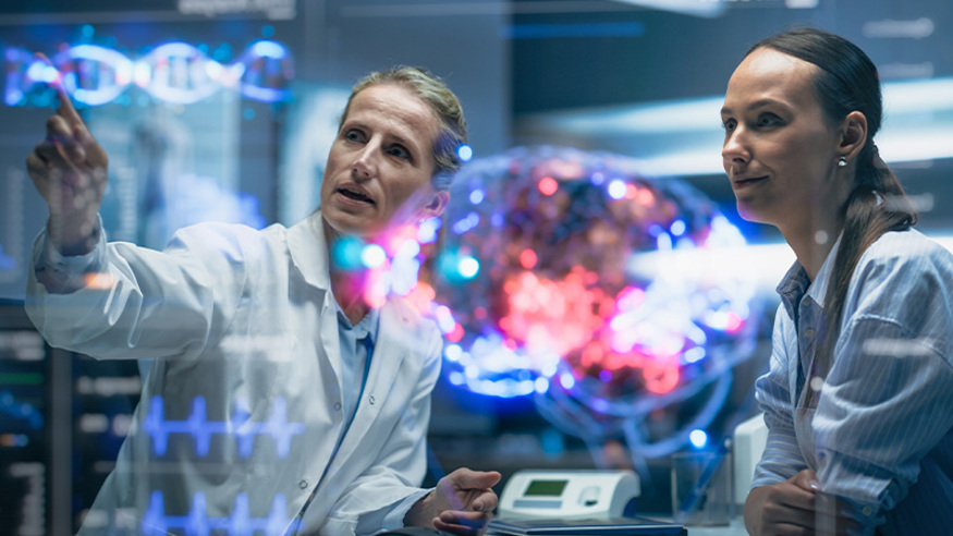 Man and woman in lab coats looking at data on a glass screen. Photo is shot from behind the glass screen.